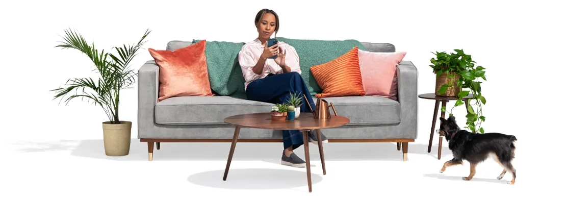 当她的狗靠近时, a young woman on a grey couch between two potted plants and colored pillows uses her smart phone to check out how much she could 保存 by switching to 国营农场.